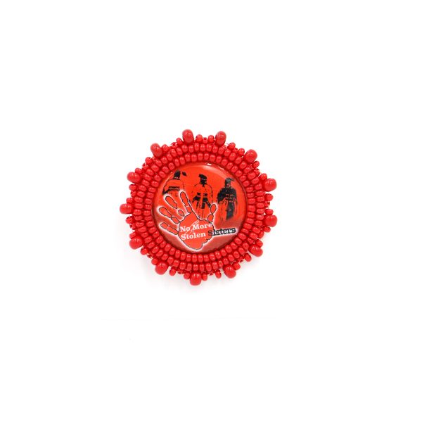 A circular object, the brooch is beaded around the edges of an acrylic pin that says "no more stolen sisters" and has imagery of people in traditional garbs and an orange handprint. The beads are red.