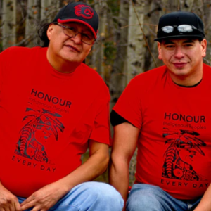 A red shirt with a graphic on it of an Indigenous man. The shirts have black text on a red background saying "Honour Indigenous Peoples Every Day." Two people are sitting side by side wearing the shirts. They are in front of a stand of birch trees.