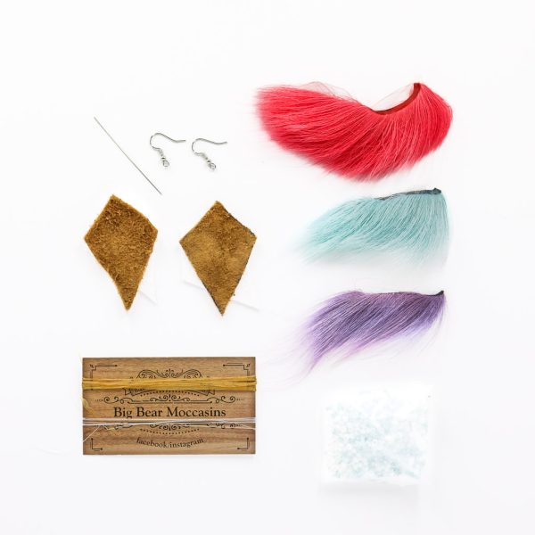 Tools and materials for beading custom earrings including a needle, two fishhook style earring findings, two pieces of hide, a business card with sinew and string wrapped around it, three tufts of animal fur in different shades, and a bag of blue and white beads.
