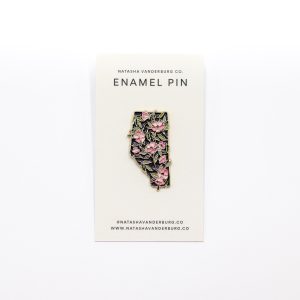 enamel pin attached to beige card, detailed enamel work in the shape of a field of wildrose flowers over an alberta shaped pin.